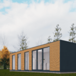 Maison modulaire moderne avec panneaux extérieurs en bois, grandes portes et fenêtres en verre, située dans une zone herbeuse avec des arbres en arrière-plan et un ciel partiellement nuageux.