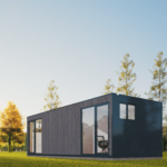 Bureau modulaire moderne avec grandes fenêtres et portes vitrées, matériaux sombres, situé dans une zone herbeuse avec des arbres en arrière-plan, ciel clair et ensoleillé, chaise de bureau visible à l'intérieur.