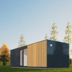 Maison modulaire moderne avec panneaux extérieurs sombres et en bois, grandes portes vitrées, petite fenêtre carrée, située dans une zone herbeuse avec arbres élancés en arrière-plan, ciel bleu dégagé.