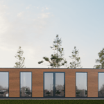 Bâtiment de bureau moderne avec extérieur en bois et grandes fenêtres en verre, situé dans un cadre naturel avec des arbres et de la verdure, intérieur visible avec chaises de bureau et bureaux.