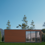 L'image montre un bureau modulaire moderne et rectangulaire avec une façade en bois et de grandes fenêtres vitrées. Le bureau est situé dans une zone herbeuse avec plusieurs arbres élancés et des montagnes en arrière-plan. Le ciel est dégagé et bleu, indiquant une journée ensoleillée. L'intérieur du bureau, visible à travers les fenêtres vitrées, semble être un espace de travail ou de réunion, avec des chaises et des tables.