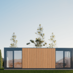 Bureau modulaire moderne avec façade en bois et grandes portes en verre, situé dans une zone herbeuse avec des arbres élancés en arrière-plan, ciel dégagé avec un léger ensoleillement, intérieur visible avec bureau et chaise.