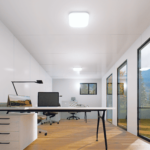Bureau moderne avec un design minimaliste, grandes fenêtres laissant entrer la lumière naturelle, deux bureaux équipés de matériel informatique, et une vue extérieure sur un paysage avec des arbres et un ciel dégagé.