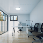 Espace de bureau modulaire moderne avec grande fenêtre et porte coulissante en verre sur le côté gauche, bureau avec ordinateur, lampe de bureau et fournitures, trois chaises de bureau ergonomiques, sol en bois poli, murs clairs.