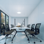 Bureau modulaire moderne avec des espaces de travail distincts, tables avec ordinateurs et chaises de bureau ergonomiques, grandes fenêtres offrant une lumière naturelle abondante, murs blancs, sol en bois clair, plantes en pot.