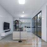 Salon moderne avec canapé clair, coussins rayés, lampe sur pied noire, télévision murale, cuisine avec armoires blanches et réfrigérateur, grandes fenêtres offrant une vue sur les arbres, sol en bois.