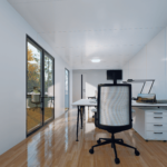 Espace de bureau moderne dans une maison modulaire avec un grand bureau, écran d'ordinateur, lampe de bureau, livres ou documents, chaise de bureau noire avec dossier en maille, murs blancs, sol en bois, grandes fenêtres offrant une vue sur des arbres aux feuilles d'automne.