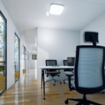 Espace de bureau moderne avec grandes fenêtres, chaises de bureau avec dossiers en filet, bureaux avec moniteurs d'ordinateur, sol en bois poli, et luminaires au plafond.