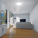 Maison modulaire moderne avec panneaux extérieurs sombres et en bois clair, grandes fenêtres et portes vitrées, cuisine avec armoires blanches et plan de travail en marbre, salon avec canapé clair et télévision à écran plat, lampe de sol, luminaire au plafond, située dans une zone herbeuse avec des arbres visibles à travers les fenêtres.