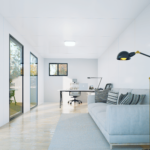 Bureau modulaire moderne avec de grandes fenêtres, un canapé de couleur claire avec des coussins à motifs, une lampe de sol noire avec un intérieur jaune, un bureau avec un ordinateur, une lampe de bureau, une chaise de bureau, un sol en bois et un tapis clair.