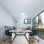 Salle de conférence moderne avec grande table blanche entourée de chaises de bureau noires sur roulettes, ornée d'une lampe de bureau, d'un calendrier et de livres, grandes fenêtres offrant une vue sur des arbres, design épuré avec murs blancs et sol en bois clair.