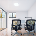 Bureau modulaire moderne avec deux chaises de bureau ergonomiques, bureau avec ordinateur portable, lampe de bureau et petits objets, grandes fenêtres sur les murs de gauche et du fond, sol en bois, murs blancs.