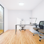 Bureau modulaire moderne avec grandes portes coulissantes en verre et panneaux extérieurs sombres, intérieur avec bureau blanc, lampe de bureau noire, ordinateur portable, tablette, livres, chaises de bureau blanches et noires, situé dans une zone herbeuse avec des arbres en arrière-plan, sous un ciel bleu et clair.