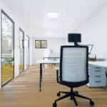 Bureau moderne et minimaliste avec postes de travail équipés, chaises de bureau noires, grandes fenêtres et vue sur un espace extérieur coloré.