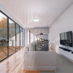 Espace de vie moderne avec canapé confortable, coussins rayés, télévision à écran plat, fenêtres vitrées du sol au plafond, terrasse en bois avec table à manger, vue sur des arbres et des montagnes, petite cuisine avec armoires blanches, bureau avec chaise, lumière naturelle, luminaire au plafond, sol en bois et tapis sous le canapé.