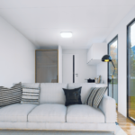 Maison modulaire moderne avec canapé gris clair, coussins décoratifs, grandes fenêtres offrant une vue sur un espace extérieur avec des arbres et des montagnes, petite cuisine avec armoires blanches et ustensiles de cuisine, lampe de sol noire, placard avec porte coulissante en verre, porte blanche menant à une autre pièce.