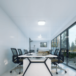 Espace de bureau modulaire moderne avec grande table centrale et plusieurs chaises de bureau, lampe de bureau, pile de livres, fournitures de bureau, grandes fenêtres laissant entrer la lumière naturelle, vue sur des arbres, murs et plafond blancs, lumières au plafond.