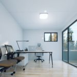 Bureau modulaire moderne avec intérieur blanc épuré, sol en bois clair, deux chaises ergonomiques, long bureau avec lampe de bureau, ordinateur portable et fournitures de bureau, grande fenêtre sur le côté droit, petite fenêtre sur le mur du fond, vue sur des arbres à l'extérieur, luminaire moderne au plafond.