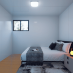 Petite chambre modulaire avec lit, literie blanche et couverture gris foncé, oreillers de différentes tailles et couleurs, table de nuit avec lampe et cadre photo, murs blancs, sol en bois, fenêtre au-dessus du lit, luminaire carré au plafond.