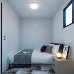 Chambre modulaire moderne avec lit, literie blanche et couverture grise, coussins noirs et blancs, livre ouvert sur le lit, petite table de nuit avec lampe et cadre photo, fenêtre laissant entrer la lumière naturelle, murs blancs, plafond blanc, porte blanche avec poignée moderne, sol avec tapis à motifs.