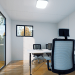 Espace de bureau moderne avec grande fenêtre, chaises de bureau avec dossiers en filet, bureau blanc avec cahier et ordinateur portable, sol en bois poli, et luminaire carré au plafond.