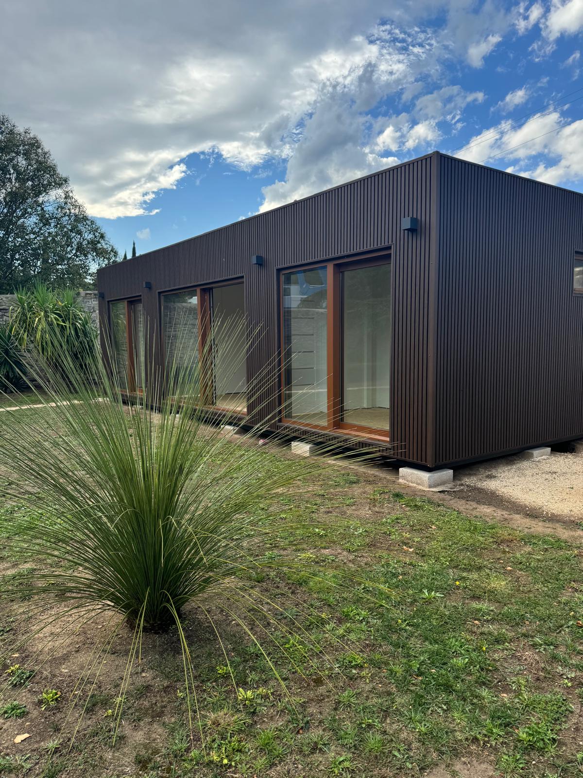 Maison modulaire moderne avec un extérieur brun foncé et des lignes verticales, de grandes portes coulissantes en verre avec des cadres en bois, légèrement surélevée sur des blocs de béton, entourée de zones herbeuses et de sol nu, avec une plante buissonnante à l'avant, sous un ciel partiellement nuageux avec des zones de ciel bleu.