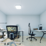 Espace de bureau moderne avec trois chaises ergonomiques et trois bureaux, chacun équipé d'une lampe de bureau, un bureau avec ordinateur portable et cadre photo, murs clairs et sol en bois, conçu pour le travail collaboratif.